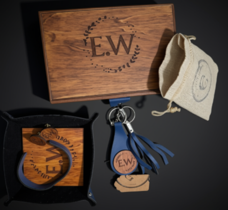 Personalized leather keychain and bracelet gift set with a wooden box engraved with 'E.W', a beige drawstring pouch, and a blue leather keychain with tassel and matching bracelet on a black background.